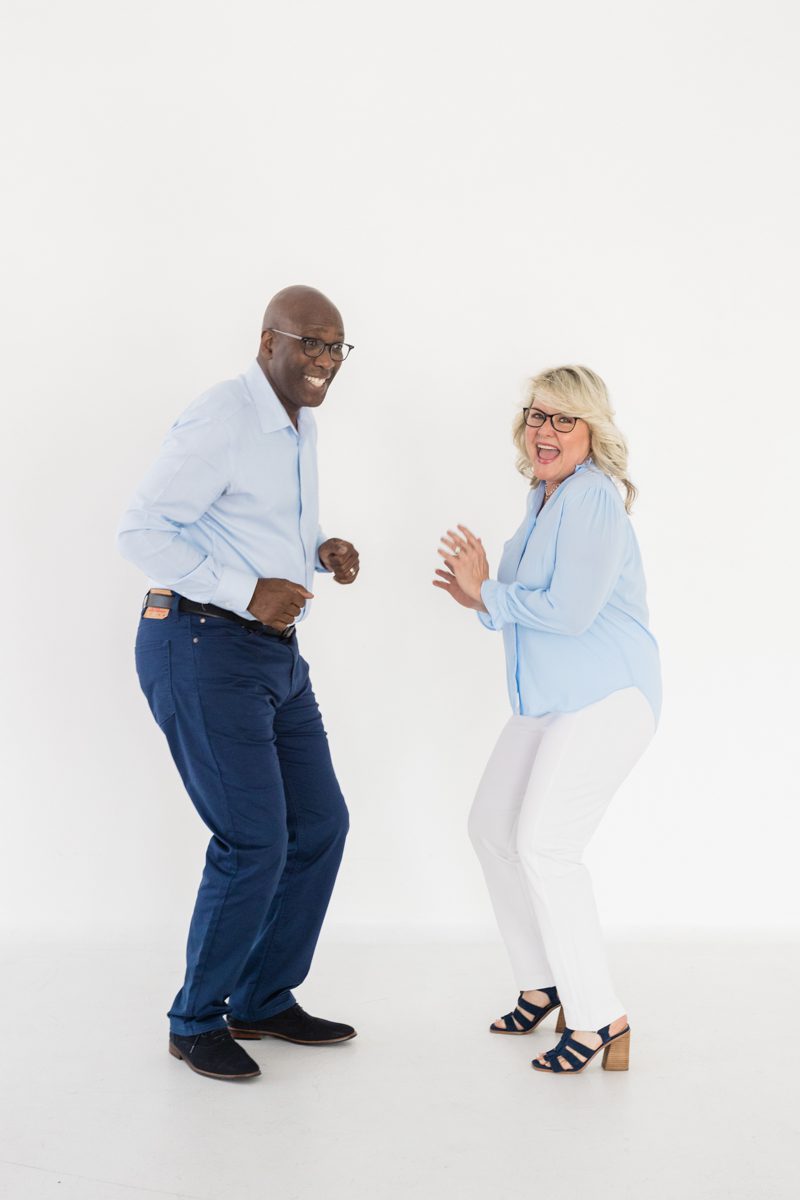 A man in a light blue dress shirt and blue jeans dances and smiles with a woman in a light blue dress shirt and white pants.