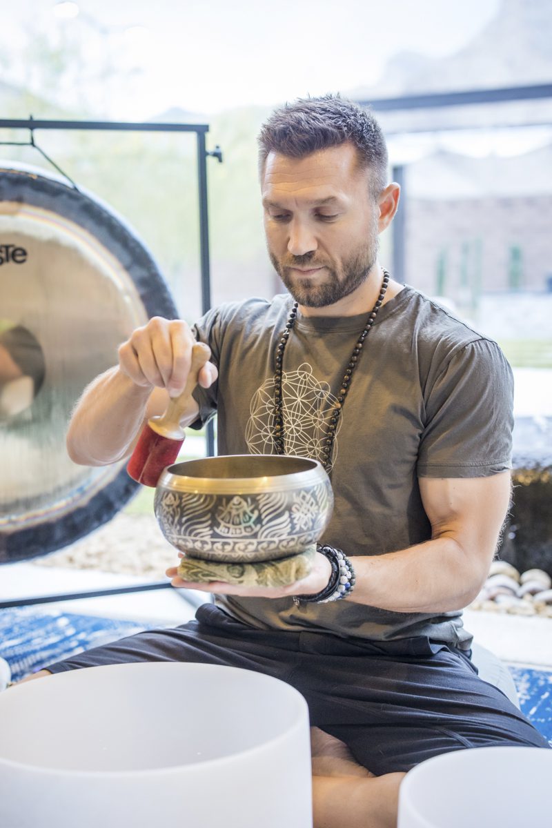 A bearded man in a light brown t-shirt plays a singing bowl made of bronze. He sits on the floor of a yoga studio with a large gong in the background.