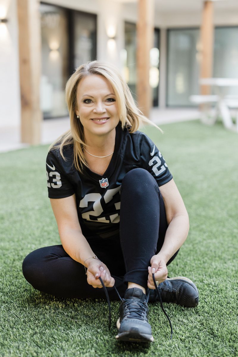 A blond-haired woman sits on artificial turf in a front yard to tie her tennis shoe. She is wearing black pants and a black NFL Las Vegas Raiders jersey.