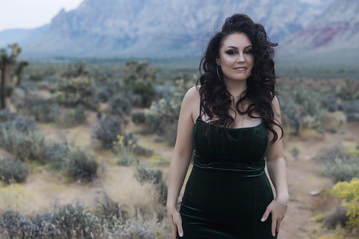 A woman with curled black hair and a green velvet dress stands amongst Joshua trees and blooming desert flowers at the Red Rock Canyon National Conservation Area.