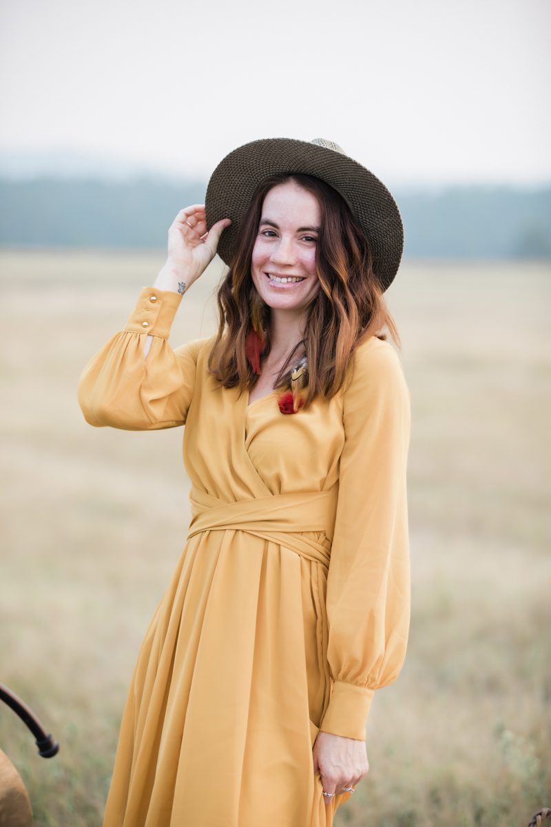 A red-haired woman in a long yellow dress tips her wide brimmed hat and smiles. She is standing in an open field. The background is blurred.