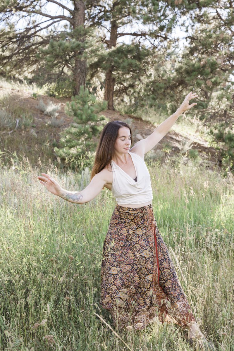 A woman in a long colorful paisley print skirt appears to be dancing in the woods. There are pine trees on the hill behind her. She stands in the knee-high grass growing in the foreground.