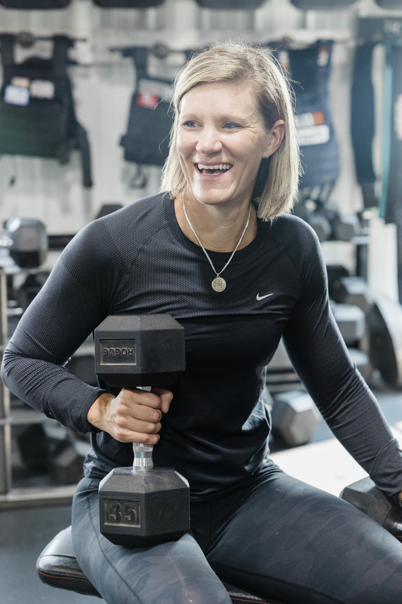 A woman in workout clothing sits on a weightlifting bench at the gym. She is resting a 35-pound dumbbell on her right thigh. She looks off camera and smiles.