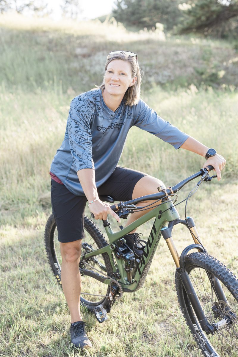 A woman straddles her green Pivot brand mountain bike. She is in a grassy field with a hill behind her. The late afternoon sun shines in from the viewer's right.