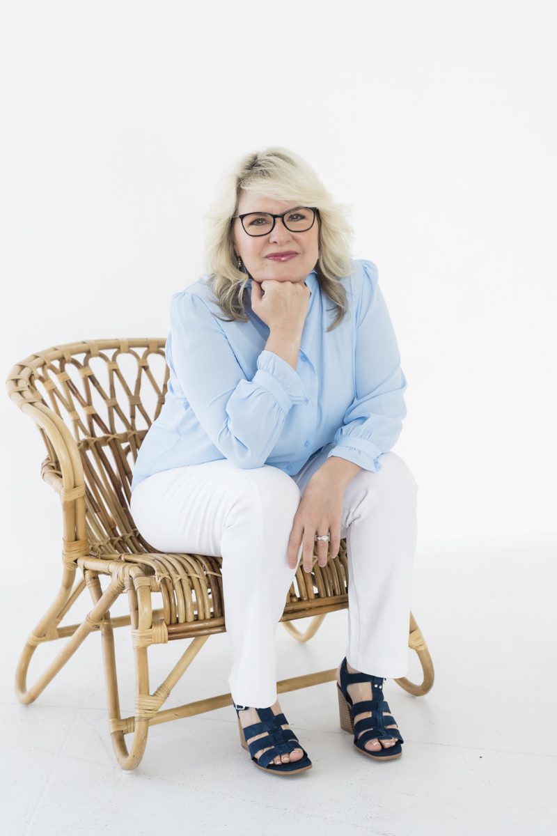 A portrait of a woman wearing glasses sitting in a curvy bamboo chair set against an all-white background. The woman leans forward and rests her chin on her fist which is positioned upright and planted on her knee.