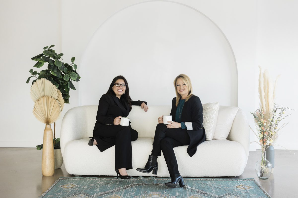 Two women in business suits sit on a white couch and each hold a white coffee mug. The rug under their feet is aqua colored. The modern looking white room is decorated with a fiddle leaf fig, trimmed dried palm leaves, pampas grass stems and budding branches.