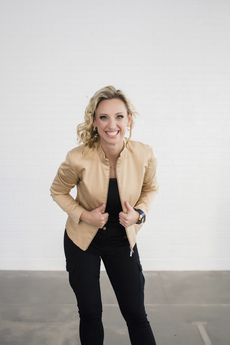 A woman in a tan leather jacket, black shirt and black pants smiles big and grips the front panels of her jacket. The wall behind her is white and the concrete floor is grey.