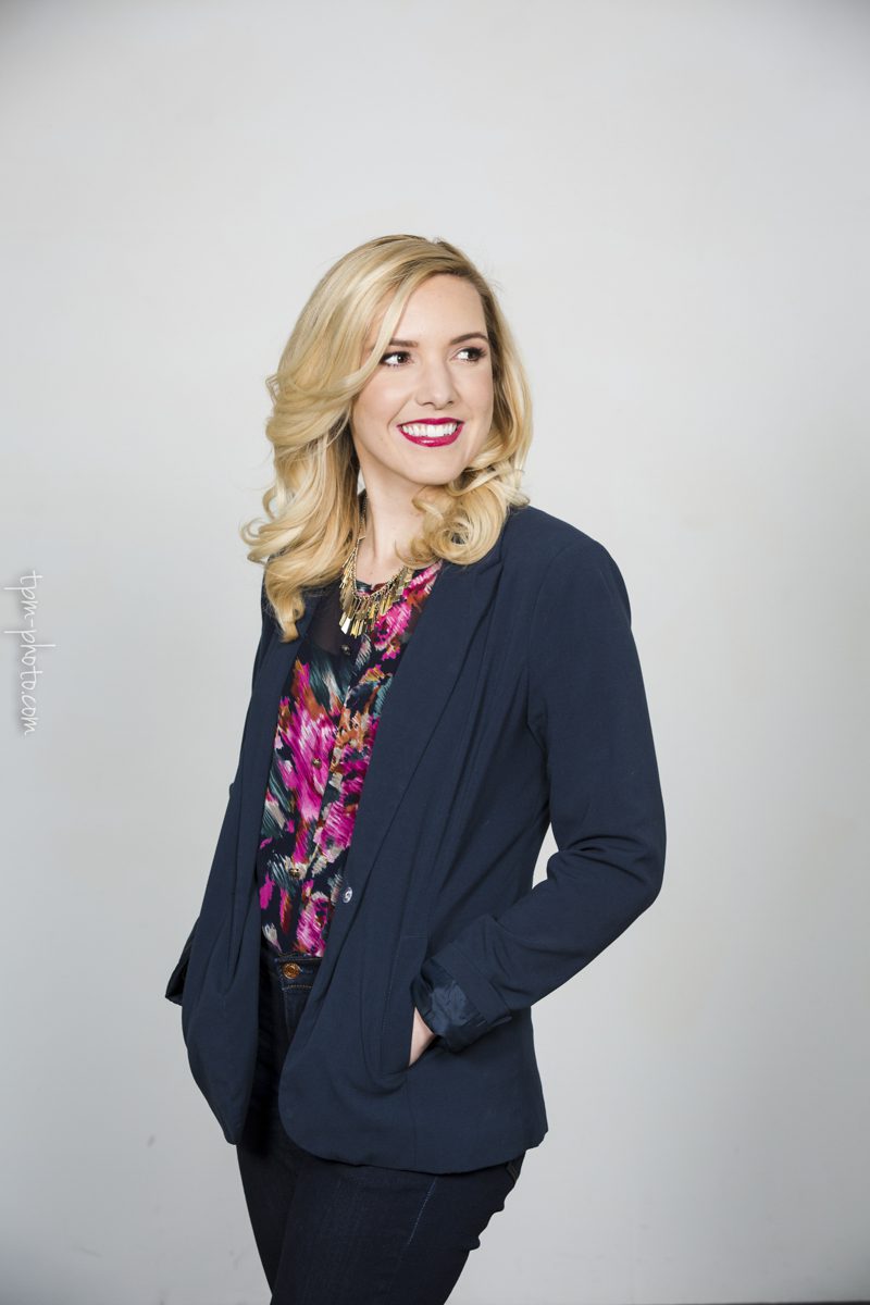 A portrait of a woman with curly long blond hair and bright red lipstick standing in a quarter turn pose and looks sideways over her left shoulder. She is wearing dark pants, a fashionable dark blue jacket and a colorful shirt.