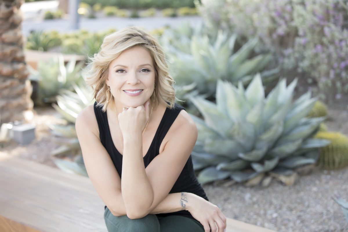 A blond-haired woman wearing a sleeveless black shirt rests her chin on her hand which is positioned upright on her knee. She sits in front of a raised flower bed which has several agave and barrel cactus growing in it.