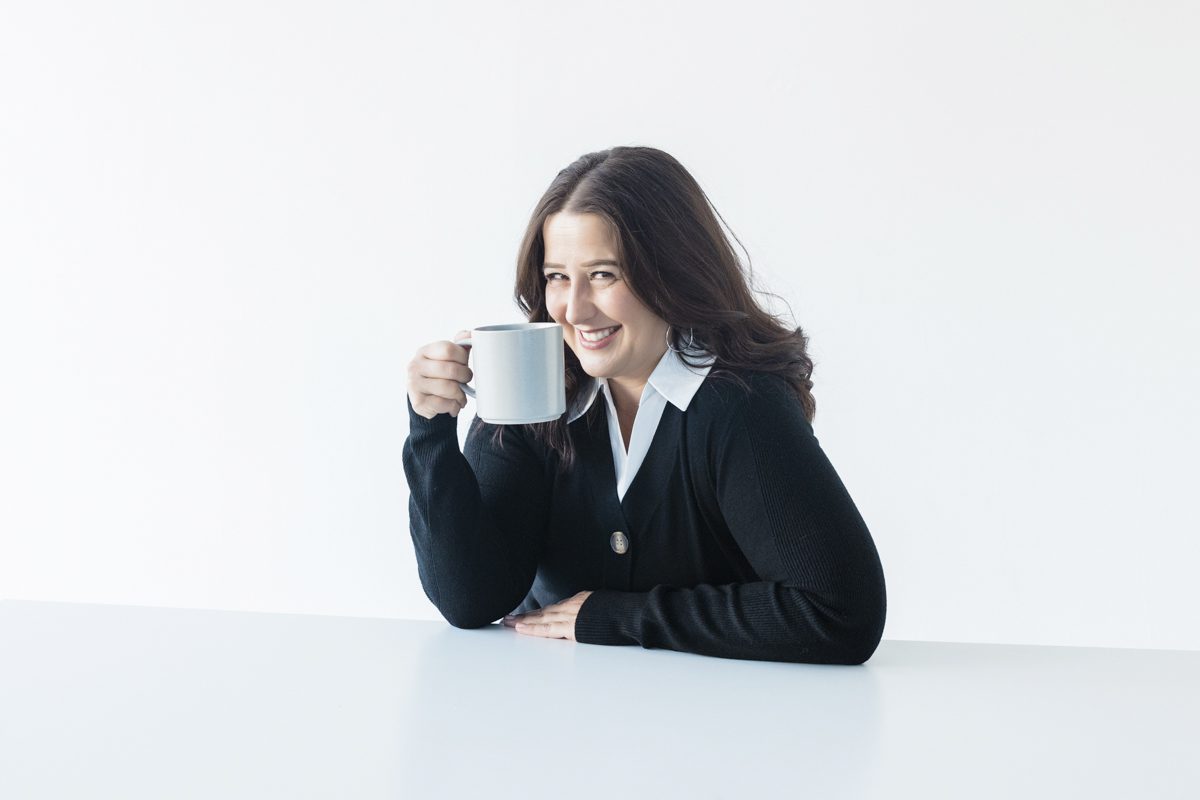 A portrait of a woman with dark hair sits behind a white table and holds a white coffee mug to her face. The background is also white. She wears a dark blazer and a white shirt with a collar that folds over her jacket.