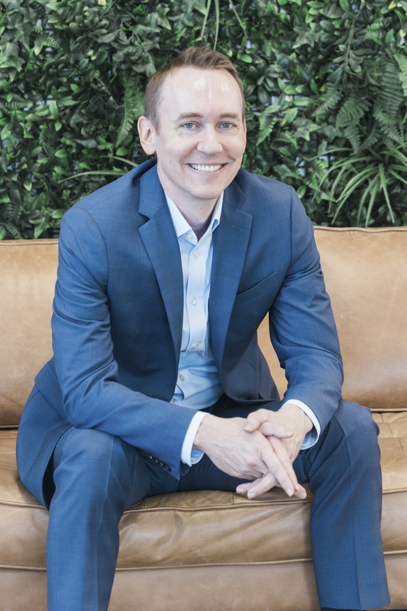 A portrait of a smiling man sitting on a tan leather couch in front of a green plant wall. The man wears a blue suit and an unbuttoned dress shirt. He leans forwards and rests his elbows on his knees and holds his hands together.
